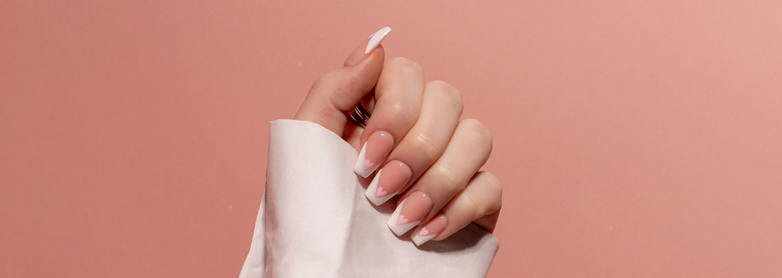 Close-up van een gemanicuurde hand met French tips en hartjes nailart op een zachte roze achtergrond, symbool voor veilige, TPO-vrije nagelverzorging.